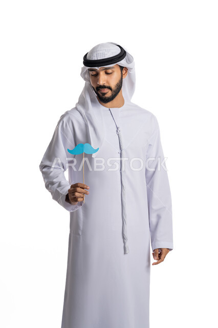 Commemoration of International Men's Day, respect and appreciation for men, the concept of strength and masculinity, a portrait of a young Arab Gulf Emirati man wearing a kandora and a ghutra, holding in his hand a piece of paper in the shape of a mustache that he looks at, a white background.