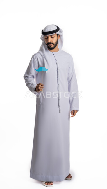 اليوم العالمي للرجل 19 نوفمبر، احترام وتقدير الرجال، مفهوم القوة والرجولة، بورتريه لشاب عربي خليجي إماراتي يرتدي الكندورة والغترة يمسك بيده قصاصة ورقية على شكل شارب يقوم بالنظر إليها، صورة لكامل الجسم، خلفية بيضاء