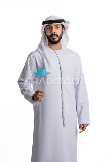 الوقوف باستقامة والنظر إلى الكاميرا بإيماءات الحزم والجدية، الاهتمام بصحة الرجال، الاحتفال باليوم العالمي للرجل، مفهوم القوة والرجولة، بورتريه لشاب عربي خليجي إماراتي يرتدي الكندورة والغترة يمسك بيده عود خشبي عليه قصاصة ورقية على شكل شارب، خلفية بيضاء