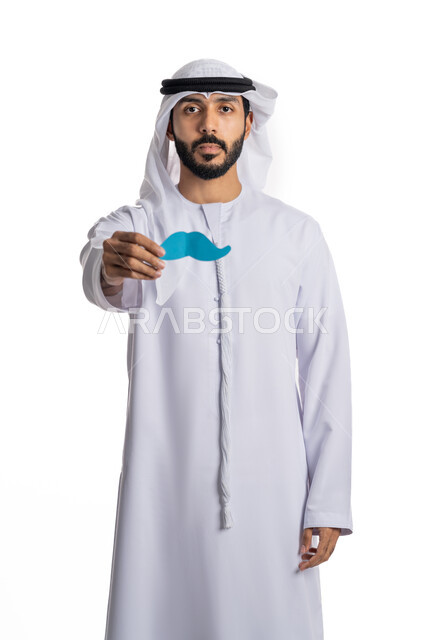 احترام وتقدير الرجال، الاحتفال باليوم العالمي للرجل، مفهوم القوة والرجولة، بورتريه لشاب عربي خليجي إماراتي يرتدي الكندورة والغترة يمسك بيده قصاصة ورقية على شكل شارب، النظر إلى الكاميرا بإيماءات الحزم والجدية، خلفية بيضاء
