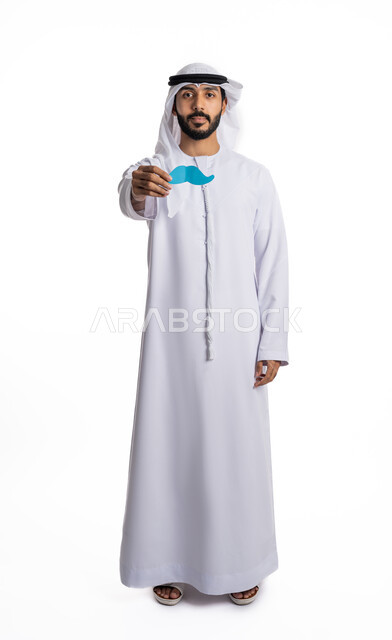 الاهتمام بصحة الرجال، الاحتفال باليوم العالمي للرجل، مفهوم القوة والرجولة، بورتريه لشاب عربي خليجي إماراتي يرتدي الكندورة والغترة يمسك بيده قصاصة ورقية على شكل شارب، النظر إلى الكاميرا بإيماءات الثقة بالنفس، صورة لكامل الجسم، خلفية بيضاء