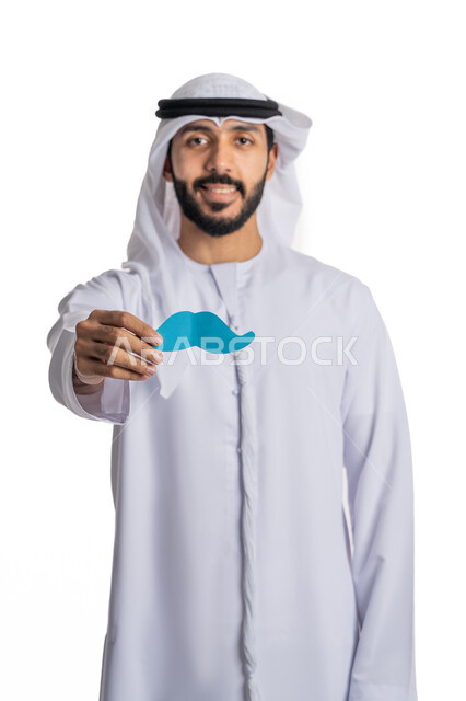 الاحتفال باليوم العالمي للرجل، مفهوم القوة والرجولة، بورتريه لشاب عربي خليجي إماراتي يرتدي الكندورة والغترة يمسك بيده قصاصة ورقية على شكل شارب، الاهتمام بصحة الرجال، النظر إلى الكاميرا بإيماءات البهجة والسرور، خلفية بيضاء