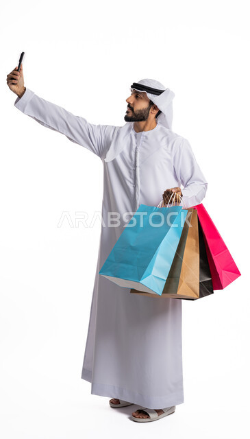 الاستفادة من العروض في شراء الحاجيات الضرورية، استلام طلبات الاونلاين، متعة التسوق واقتناء الهدايا، بورتريه لرجل عربي خليجي إماراتي يرتدي الكندورة والغترة يمسك بيده أكياس ورقية يقوم بالتقاط صورة سيلفي باستخدام الهاتف المحمول، صورة لكامل الجسم، خلفية بيضاء
