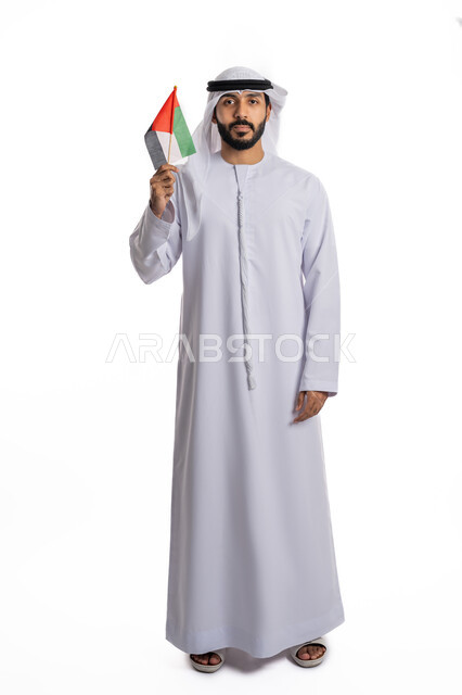 مفهوم حب الوطن والانتماء إليه، بورتريه لرجل عربي خليجي إماراتي يرتدي الكندورة والغترة يرفع علم الإمارات المتحدة، الاحتفال باليوم الوطني، إحياء ذكرى استقلال البلاد، الوقوف باستقامة والنظر إلى الكاميرا بإيماءات الاعتزاز والفخر، صورة لكامل الجسم، خلفية بيضاء