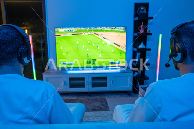 المتعة والتسلية بالفيديو جيمز برفقة الأصدقاء, المشاركة والمنافسة بالألعاب التقنية الحديثة والمتطورة, صورة من الخلف لرجلين عربيين خليجيين اماراتيين يرتديان الكندورة الاماراتية وسماعة الهيدفون يستمتعان بقضاء أوقات مسلية, العالم الرقمي الترفيهي