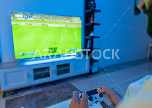 استخدام الأجهزة والتقنيات الحديثة والمتطورة, دمج المتعة والتسلية مع العالم الالكتروني, صورة مقربة من الخلف لرجل عربي خليجي إماراتي يرتدي الكندورة يمسك بيده عصا التحكم, العالم الرقمي الترفيهي, قضاء أوقات مسلية بألعاب الفيديو