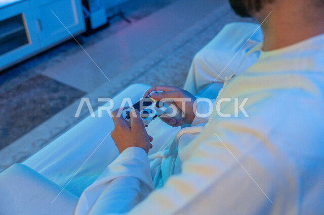Using modern and advanced devices and technologies, fun and entertainment with video games, interaction and integration with entertainment electronic games, a picture from the side of an Arab Gulf Emirati young man wearing an Emirati kandora holding a joystick in his hand, spending fun times.