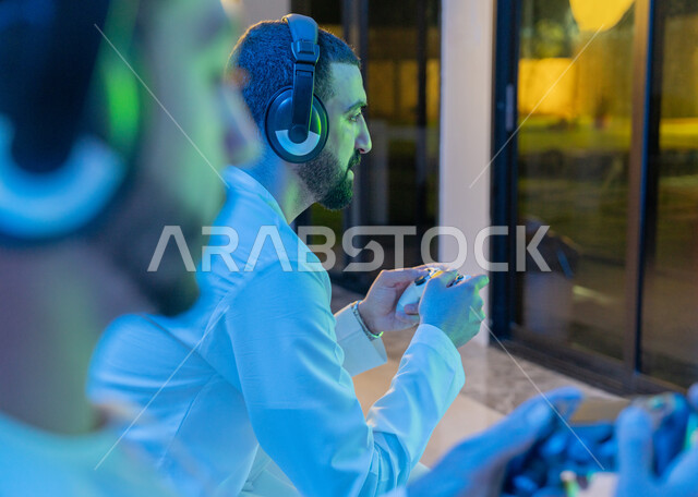العالم الرقمي الترفيهي, المتعة والتسلية بالفيديو جيمز برفقة الأصدقاء, المشاركة والمنافسة بالألعاب التقنية الحديثة والمتطورة, صورة مقربة من الجانب لرجلين عربيين خليجيين اماراتيين يرتديان الكندورة الاماراتية وسماعة الهيدفون يمسكان عصا التحكم