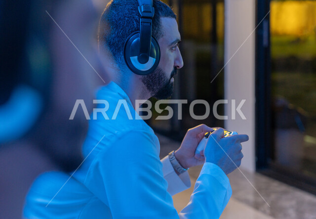 المشاركة والمنافسة بالألعاب التقنية الحديثة والمتطورة, صورة مقربة من الجانب لرجلين عربيين خليجيين اماراتيين يرتديان الكندورة الاماراتية وسماعة الهيدفون يمسكان عصا التحكم, العالم الرقمي الترفيهي, المتعة والتسلية بالفيديو جيمز برفقة الأصدقاء