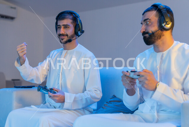 رفع اليدين بحماس لتحقيق الفوز والانتصار, الاندماج والتفاعل بألعاب الفيديو, قضاء أوقات ممتعه برفقة الأصدقاء, أنشطة شبابية ترفيهية, شابان عربيان خليجيان إماراتييان يرتديان الكندورة الاماراتية يمسكان عصا التحكم ويلعبان بالألعاب الالكترونية