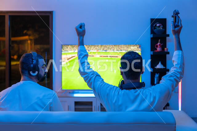 Integration, interaction and enthusiasm in the modern and advanced electronic digital world, a picture from the back of two young Arab Gulf Emirati men wearing the Emirati kandora and a headphone competing in video games, raising hands with gestures of victory and triumph, fun and entertaining entertainment experiences.