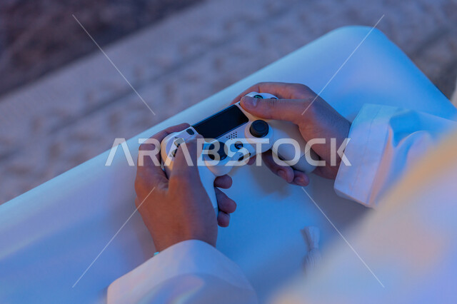 دمج المتعة والتسلية مع العالم الالكتروني, صورة مقربة ليدين رجل عربي خليجي إماراتي يرتدي الكندورة يمسك بيده عصا التحكم, العالم الرقمي الترفيهي, قضاء أوقات مسلية بألعاب الفيديو, استخدام الأجهزة والتقنيات الحديثة والمتطورة