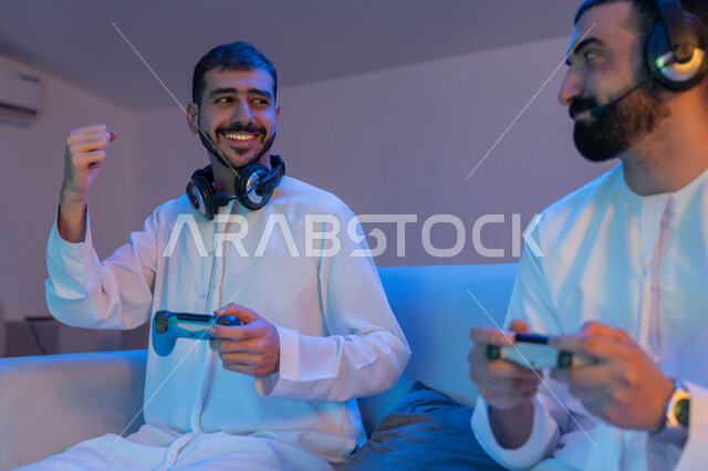 قضاء أوقات ممتعه برفقة الأصدقاء, أنشطة شبابية ترفيهية, شابان عربيان خليجيان إماراتييان يرتديان الكندورة الاماراتية يمسكان عصا التحكم ويلعبان بالألعاب الالكترونية, رفع اليدين بإيماءات الاندماج والتفاعل والحماس بألعاب الفيديو
