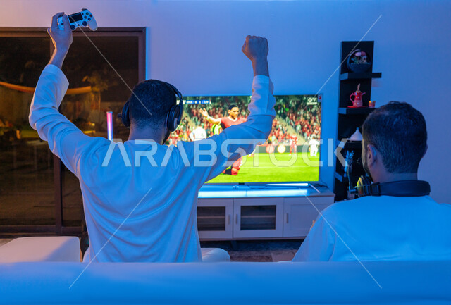 رفع اليدين بإيماءات الفوز والانتصار, تجارب ترفيهية ممتعة ومسلية, الاندماج والتفاعل والحماس بالعالم الرقمي الالكتروني الحديث والمتطور, صورة من الخلف لشابين عربيين خليجيين إماراتيين يرتديان الكندورة الاماراتية وسماعة الهيدفون يقومان بالمنافسة بالفيديو جيمز