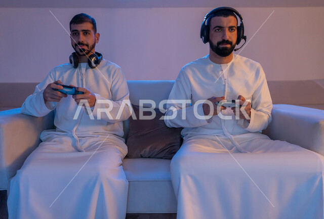 قضاء أوقات ترفيهية حماسية شبابية ممتعة, التركيز والتفاعل والاندماج بالألعاب الالكترونية, شابان عربيان خليجيان اماراتيان يرتديان الكندورة الاماراتية وسماعة الهيدفون يمسكان عصا التحكم ويقومان بالتنافس بالفيديو جيمز