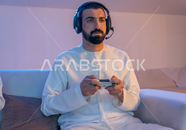 المتعة والتسلية بالفيديو جيمز, العالم الرقمي الترفيهي، شاب عربي خليجي إماراتي يرتدي الكندورة الاماراتية وسماعة الهيدفون يمسك بيده عصا التحكم بتركيز واندماج, الاستمتاع بتجربة ترفيهية ممتعة ومثيرة في الألعاب الالكترونية والأجهزة الحديثة