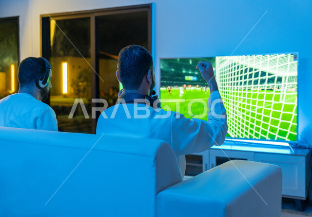 Enjoyable and entertaining entertainment experiences, integration, interaction and enthusiasm in the modern and advanced electronic digital world, a picture from the back of two Arab Gulf Emirati young men wearing the Emirati kandora and a headphone competing in video games, raising hands with victory and victory gestures