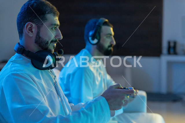 التركيز والتفاعل والاندماج, الألعاب الالكترونية الترفيهية الممتعة والمسلية برفقة الأصدقاء, صورة مقربة من الجانب لشابين عربيين خليجيين إماراتيين يرتديان الكندورة الاماراتية وسماعة الهيدفون يمسكان عصا التحكم ويقومان بالتنافس بالفيديو جيمز