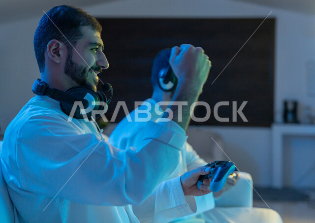 Raising hands with gestures of enthusiasm, interaction and integration, fun and entertaining electronic games with friends, close-up photo from the side of two Arab Gulf Emirati young men wearing Emirati kandura and headphones holding a joystick and competing in video games