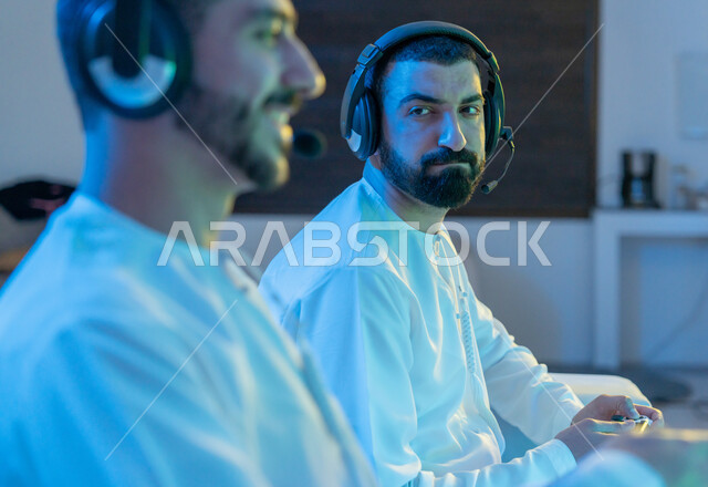 الألعاب الالكترونية الترفيهية الممتعة والمسلية برفقة الأصدقاء, صورة مقربة من الجانب لشابيين عربيين خليجيين إماراتيين يرتديان الكندورة الاماراتية وسماعة الهيدفون يمسكان عصا التحكم ويقومان بالتنافس بالفيديو جيمز, استخدام الأجهزة والتقنيات الحديثة والمتطورة