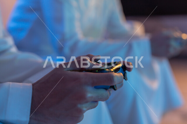 المشاركة والمنافسة بالألعاب التقنية الحديثة والمتطورة, الاستمتاع بقضاء الوقت الممتع برفقة الأصدقاء, صورة مقربة ليد رجلين عربيين خليجيين اماراتيين يرتديان الكندورة الاماراتية يمسكان عصا التحكم بتركيز, العالم الرقمي الترفيهي, المتعة والتسلية بالفيديو جيمز