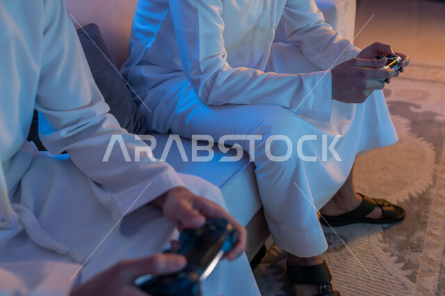 المشاركة والمنافسة بالألعاب التقنية الحديثة والمتطورة, الاستمتاع بقضاء الوقت الممتع برفقة الأصدقاء, رجلان عربيان خليجيان اماراتيان يرتديان الكندورة الاماراتية يمسكان عصا التحكم بتركيز, العالم الرقمي الترفيهي, المتعة والتسلية بالفيديو جيمز