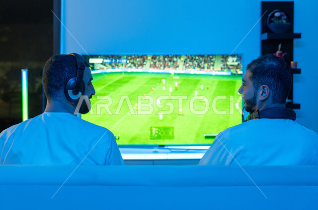 Using modern and advanced devices and technologies, fun, enthusiastic youth activities, exciting digital entertainment experiences, a picture from behind of two young Arab Gulf and Emirati men wearing the Emirati kandora, playing video games with immersion, interaction, and enthusiasm.