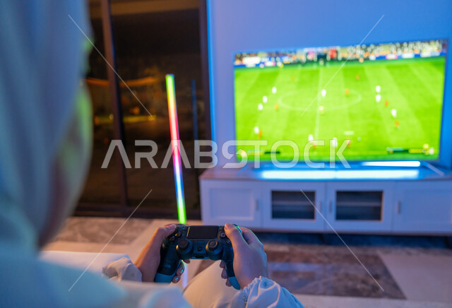 قضاء أوقات ترفيهية ممتعة في المنزل, صورة مقربة من الخلف لشابة سعودية تمسك بيدها عصا التحكم, إمرأة عربية خليجية إماراتية ترتدي العباءة البيضاء تقوم باللعب والتسلية بالفيديو جيمز, استخدام أجهزة تقنية الكترونية حديثة ومتطورة