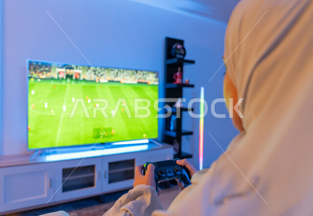 Using modern and advanced electronic technical devices, spending enjoyable entertainment times at home, a close-up photo from the back of a Saudi young woman holding a joystick in her hand, an Arab Gulf Emirati woman wearing a white abaya playing and having fun with video games