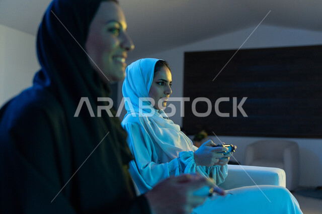 اللعب والمشاركة وقضاء وقت ممتع في المنزل, صورة من الجانب لشابة سعودية مبتسمة تمسك بيدها عصا التحكم بفرح, إمرأة عربية خليجية اماراتية ترتدي العباءة البيضاء تجلس بإيماءات التركيز والاندماج, المتعة والتسلية بألعاب الفيديو, تجارب رقمية ترفيهية