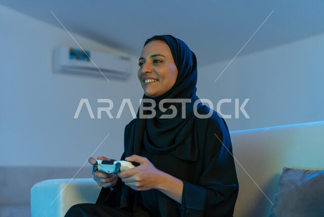 Using modern and advanced devices and technologies, fun and entertainment with video games, interaction and integration with entertainment electronic games, a close-up image of an Arab Gulf Emirati woman wearing a black abaya, holding a joystick in her hand enthusiastically, a smiling young Saudi woman enjoying a good time.