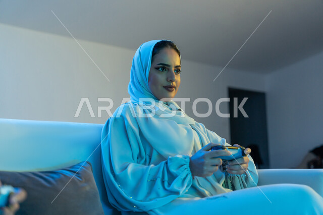 المتعة والتسلية بالفيديو جيمز, استخدام الأجهزة والتقنيات الحديثة والمتطورة, إمرأة عربية خليجية اماراتية ترتدي العباءة البيضاء تمسك بيدها عصا التحكم بتركيز واندماج, شابة سعودية تستمتع بقضاء وقت ممتع بالألعاب الالكترونية الترفيهية