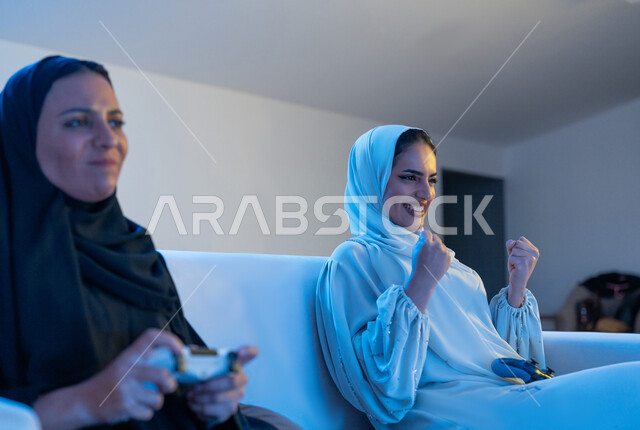 قضاء أوقات ممتعه برفقة الأصدقاء, أنشطة شبابية ترفيهية, صورة مقربة لشابة سعودية تمسك بيدها عصا التحكم بتعابير الخسارة والحزن, إمرأة عربية خليجية اماراتية ترتدي العباءة البيضاء ترفع يديها بإيماءات الاندماج والتفاعل والحماس بألعاب الفيديو