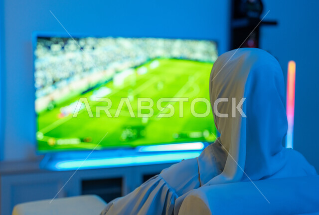 Using modern and advanced electronic technical devices, spending enjoyable recreational times at home, a close-up photo from the back of a Saudi young woman sitting with concentration and concentration, an Arab Gulf Emirati woman wearing a white abaya playing and having fun with video games