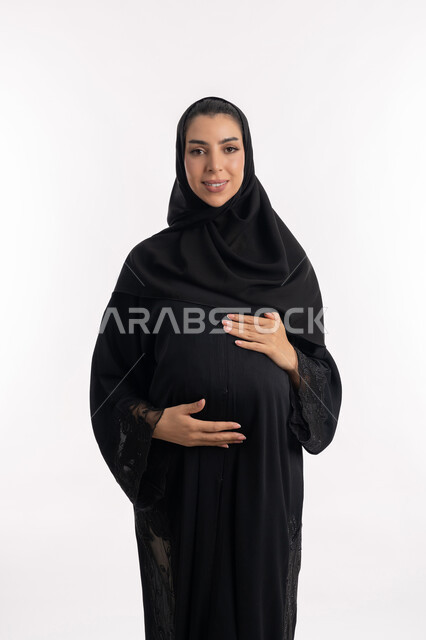 تعابير الفرح والسرور، مفهوم الحمل والانجاب، إمرأة عربية خليجية سعودية تضع يدها على بطنها، الحفاظ على صحتها و صحة طفلها، بورتريه مقرب لشابة إماراتية ترتدي العباءة السوداء  تنتظر قدوم مولودها، النظر إلى الكاميرا بإيماءات الحماس، الاهتمام بصحة الجنين، خلفية بيضاء