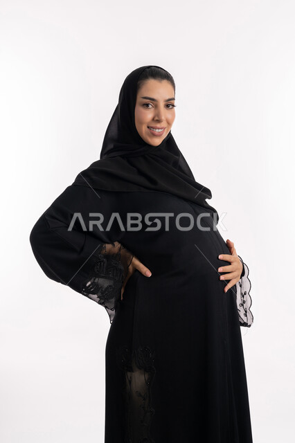 مفهوم الحمل والانجاب،  النظر إلى الكاميرا بإيماءات الحماس، إمرأة عربية خليجية سعودية تضع يدها على بطنها، الحفاظ على صحتها و صحة طفلها، الاهتمام بصحة الجنين، بورتريه مقرب لشابة إماراتية ترتدي العباءة السوداء  تنتظر قدوم مولودها، خلفية بيضاء