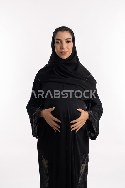 النظر إلى الكاميرا بإيماءات الحماس، إمرأة عربية خليجية سعودية تضع يدها على بطنها، الحفاظ على صحتها و صحة طفلها، الاهتمام بصحة الجنين، مفهوم الحمل والانجاب، بورتريه مقرب لشابة إماراتية ترتدي العباءة السوداء  تنتظر قدوم مولودها، خلفية بيضاء