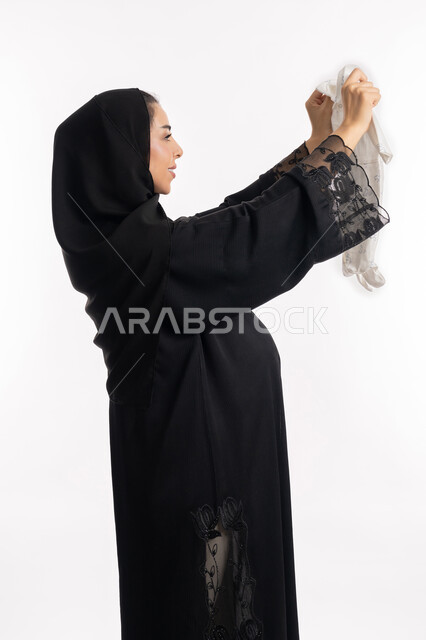 النظر إلى ملابس نوم للأطفال الرضع بإيماءات الحماس، مفهوم الحمل والانجاب، الاهتمام بصحة الجنين، الحفاظ على صحتها و صحة طفلها، إمرأة عربية خليجية سعودية تمسك بيدها افرول، بورتريه مقرب لشابة إماراتية ترتدي العباءة السوداء تنتظر قدوم مولودها، خلفية بيضاء