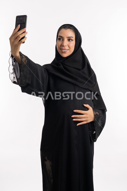 إلتقاط صورة سيلفي تذكارية عبر الجوال، توثيق اللحظات السعيدة، الحفاظ على صحتها و صحة طفلها، الاهتمام بصحة الجنين، مفهوم الحمل والانجاب، إمرأة عربية خليجية سعودية تضع يدها على بطنها، بورتريه مقرب لشابة إماراتية تمسك بيدها الهاتف المحمول، خلفية بيضاء