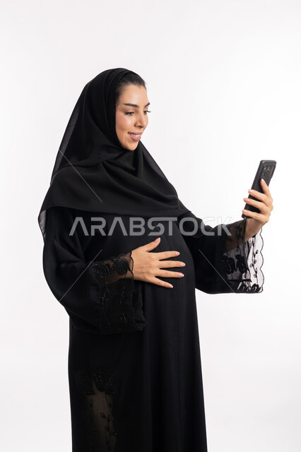 التواصل مع العائلة والأصدقاء باستخدام الهاتف المحمول، الحفاظ على صحتها و صحة طفلها، الاهتمام بصحة الجنين، مفهوم الحمل والانجاب، إمرأة عربية خليجية سعودية تضع يدها على بطنها، بورتريه مقرب لشابة إماراتية تقوم بإجراء مكالمة فيديو، خلفية بيضاء