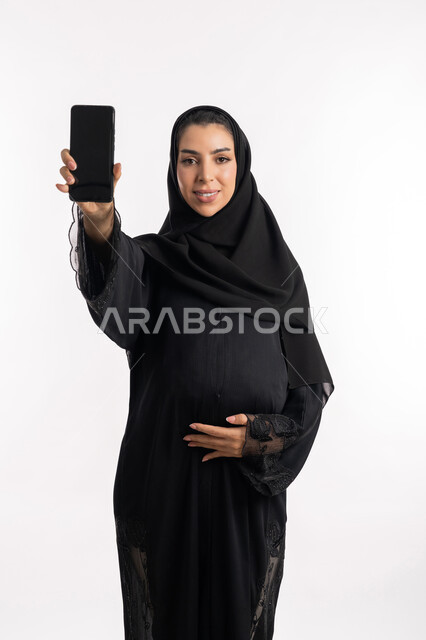 عرض شاشة سوداء فارغة عبر الهانف المحمول، الحفاظ على صحتها و صحة طفلها، الاهتمام بصحة الجنين، مفهوم الحمل والانجاب، إمرأة عربية خليجية سعودية تضع يدها على بطنها، بورتريه مقرب لشابة إماراتية ترتدي العباءة السوداء تنظر إلى الكاميرا بإيماءات الحماس، خلفية بيضاء