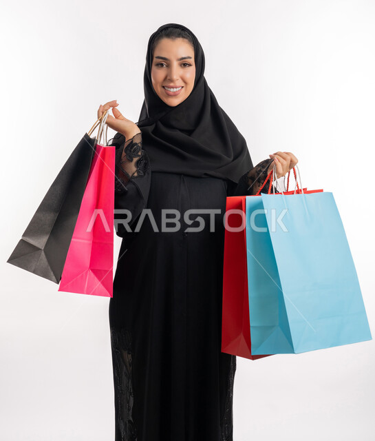 تعابير السعادة بالتسوق الممتع واقتناء المستلزمات، الاستفادة من العروض والتخفيضات الموسمية، مفهوم الحمل والانجاب،  إمرأة عربية خليجية سعودية محجبة تحمل بيدها أكياس التسوق الملونة، بورتريه مقرب لشابة إماراتية تنظر إلى الكاميرا بإيماءات السرور، خلفية بيضاء
