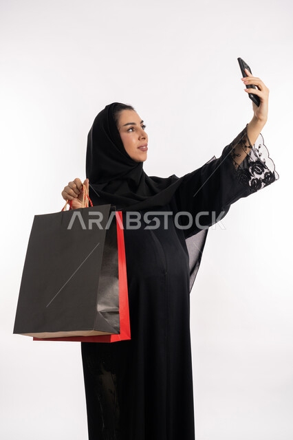 التقاط صورة سيلفي تذكارية، الاستفادة من العروض الموسمية والتخفيضات، مفهوم الحمل والانجاب،  إمرأة عربية خليجية سعودية محجبة  تحمل بيدها أكياس التسوق الملونة، بورتريه مقرب لشابة إماراتية ترتدي  العباءة السوداء تمسك بيدها الهاتف المحمول، خلفية بيضاء