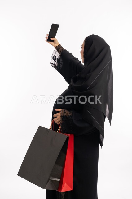 الاستفادة من العروض الموسمية والتخفيضات، مفهوم الحمل والانجاب،  إمرأة عربية خليجية سعودية  تحمل بيدها أكياس التسوق الملونة، بورتريه مقرب من الخلف لشابة إماراتية ترتدي  العباءة السوداء تمسك بيدها الهاتف المحمول تقوم بالتقاط صورة سيلفي تذكارية، خلفية بيضاء