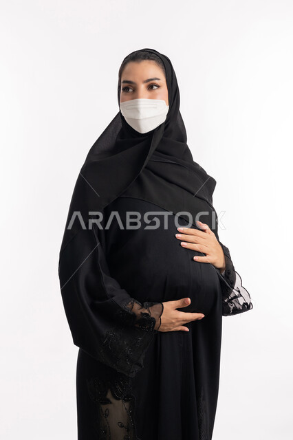 ارتداء الكمامة للوقاية من العدوى، الالتزام بالاحترازات الوقائيه ضد كوفيد 19، الاهتمام بصحة الجنين، الحفاظ على صحتها و صحة الجنين، إمرأة عربية خليجية سعودية تضع يدها على بطنها، بورتريه مقرب لشابة إماراتية ترتدي القناع الوقائي تنظر إلى مكان ما، خلفية بيضاء