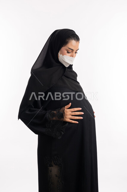 مفهوم الحمل والانجاب، الالتزام بالاحترازات الوقائيه ضد كوفيد 19، الاهتمام بصحة الجنين، ارتداء الكمامة للوقاية من العدوى، إمرأة عربية خليجية سعودية تضع يدها على بطنها، بورتريه مقرب من الجانب لشابة إماراتية ترتدي عباءة سوداء والقناع الوقائي، خلفية بيضاء