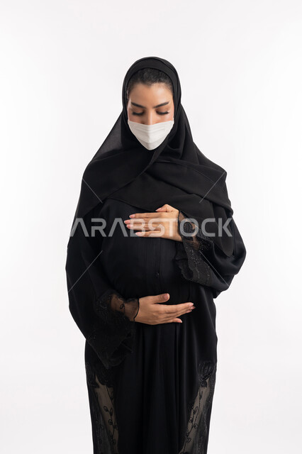 الالتزام بالاحترازات الوقائيه ضد كوفيد 19، الاهتمام بصحة الجنين، الحفاظ على صحتها و صحة الجنين، ارتداء الكمامة للوقاية من العدوى، إمرأة عربية خليجية سعودية تضع يدها على بطنها، بورتريه مقرب لشابة إماراتية ترتدي القناع الوقائي تنظر إلى الكاميرا، خلفية بيضاء