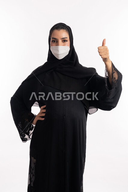 القبول والرضا والموافقة، الالتزام بالاحترازات الوقائيه ضد كوفيد 19، الاهتمام بصحة الجنين،  إمرأة حامل عربية خليجية سعودية تشير بإصبع الإبهام للأعلى، بورتريه مقرب من الجانب لشابة إماراتية ترتدي عباءة سوداء والقناع الوقائي تنظر إلى الكاميرا، خلفية بيضاء
