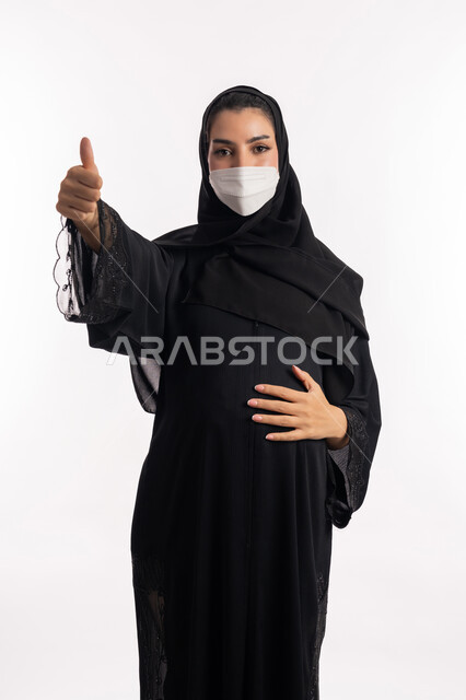 تعابير الإعجاب والرضى والقبول، الالتزام بالاحترازات الوقائيه ضد كوفيد 19، الاهتمام بصحة الجنين،  إمرأة حامل عربية خليجية سعودية تشير بإصبع الإبهام للأعلى، بورتريه مقرب من الجانب لشابة إماراتية ترتدي عباءة سوداء والقناع الوقائي تنظر إلى الكاميرا، خلفية بيضاء