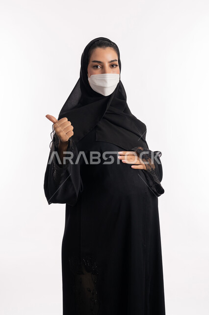 The concept of acceptance, satisfaction and approval, adherence to preventive precautions against Covid 19, concern for the health of the fetus, a pregnant Saudi Gulf Arab woman pointing with her thumb up, a close-up portrait from the side of a young Emirati woman wearing a black abaya and a protective mask looking at the camera, white background.